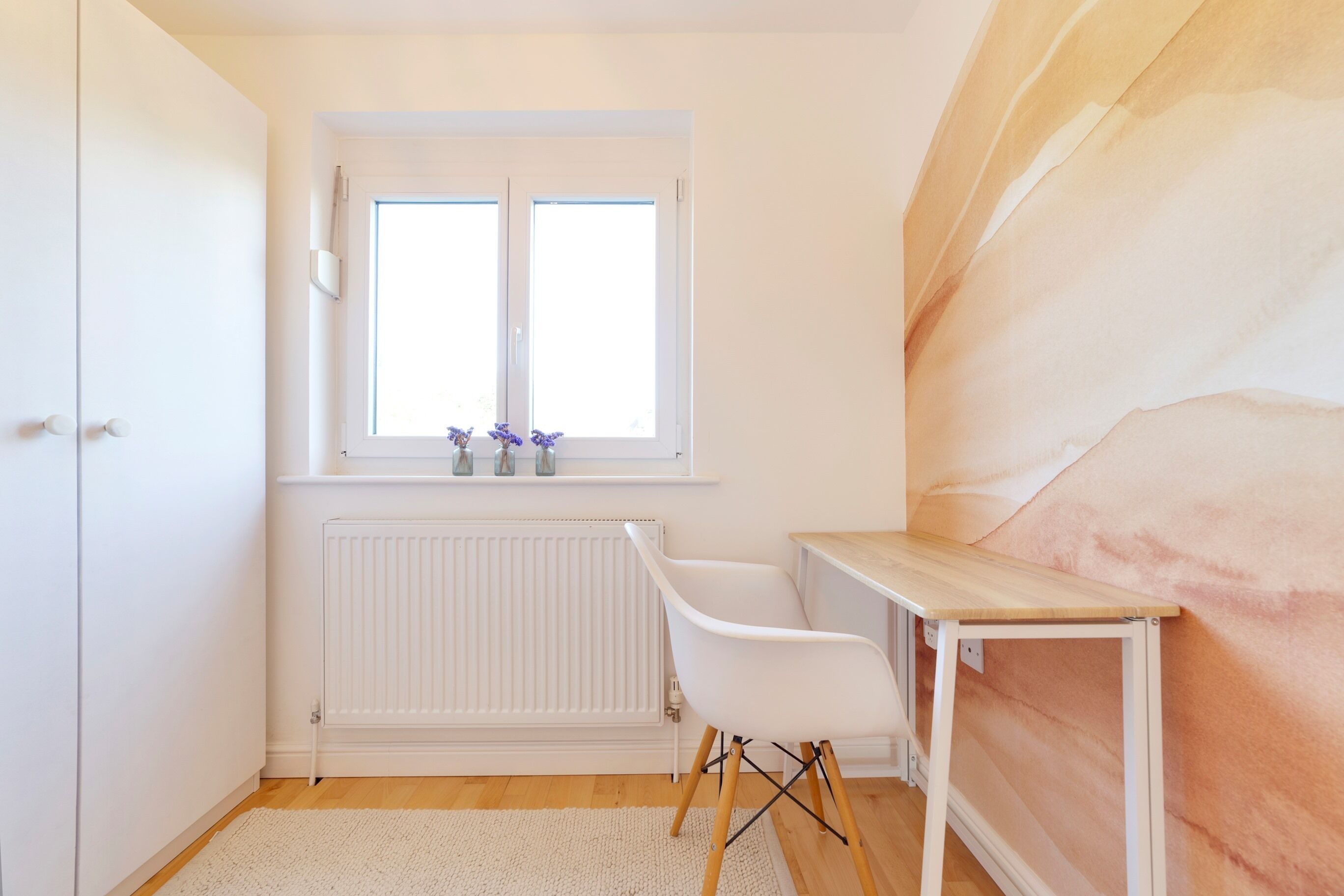 Ein Zimmer mit weißem Heizkörper, einem Fenster mit Blick und einem Holztisch sowie einem weißen Sessel.
