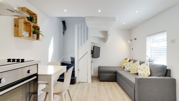 A modern kitchen with a built-in stove, a dining area with a white table and chairs, a gray sofa with patterned cushions, and a staircase leading to another level. - Pass the Keys | 19th Century Barn with Free Parking (Histon)