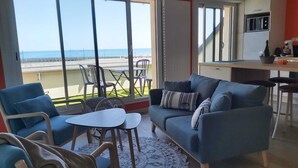 A modern living room with a sofa, chairs, and a small table. A kitchen area with a microwave is visible. Large windows offer a view of the beach.