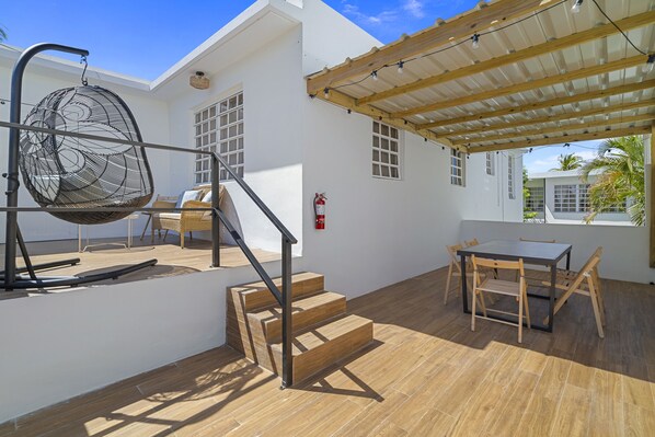 A modern outdoor patio with a wooden table and chairs, a hanging chair, and a staircase leading to an upper level.