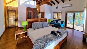 Une chambre avec un plafond en bois, un lit avec une literie à motifs, une table de chevet avec une lampe, un canapé bleu et une fenêtre donnant sur un beau paysage.