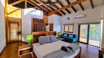 A bedroom with a wooden ceiling, a bed with patterned bedding, a nightstand with a lamp, a blue sofa, and a window with a view.