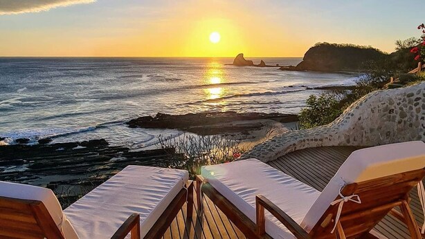 Une terrasse en bois avec deux chaises longues donnant sur un coucher de soleil sur l’océan.