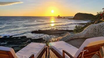 A wooden deck with two lounge chairs facing a sunset over the ocean.