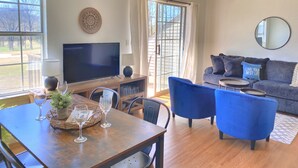 A modern living room with a flat-screen TV, a dining table with chairs, a sofa, and a large window offering a view of the outdoors. - Gorgeous and Bright, Lake Marina, Fish, Golf View! (Branson)