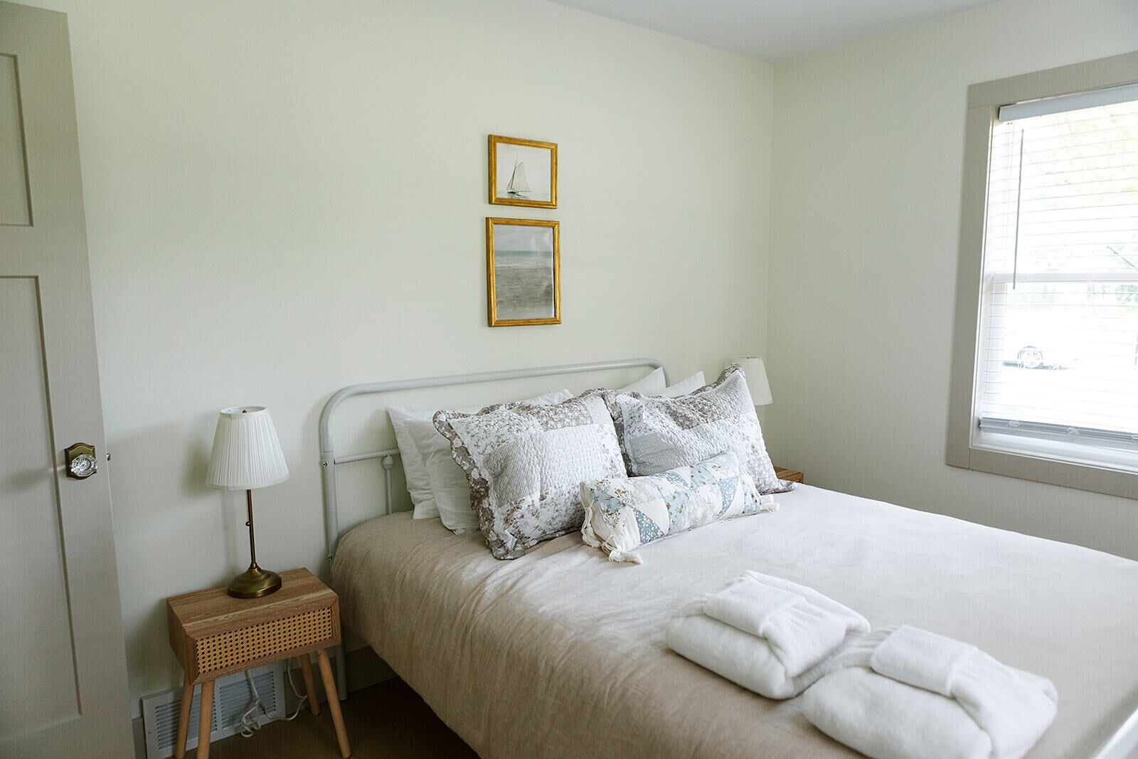 A neatly made bed with a white duvet and multiple pillows, a bedside lamp, and a window with blinds.