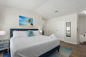 Quarto com cama grande, mesa de cabeceira, luminária, quadro de uma praia, porta e espelho.