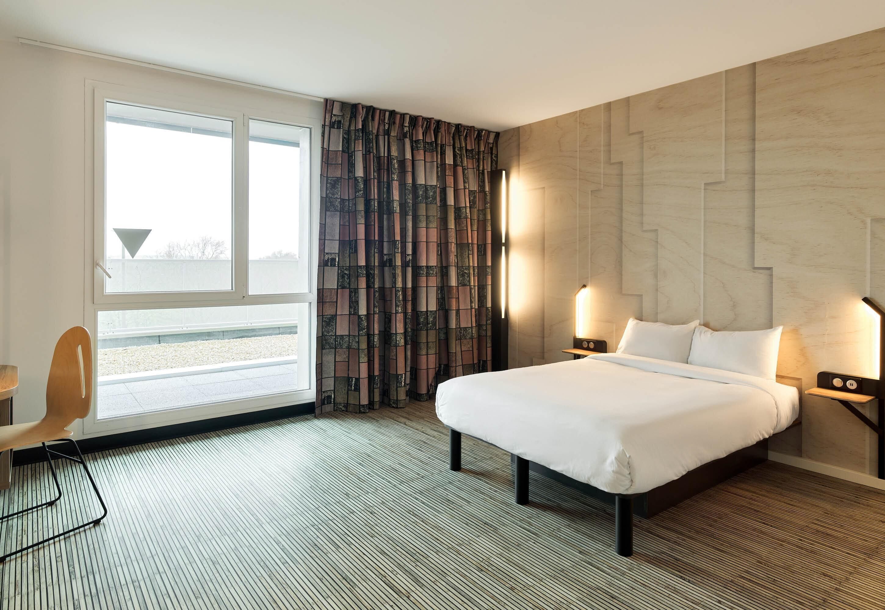 A hotel room with a large bed, a chair, a desk, a window with curtains, and a wall-mounted light fixture.