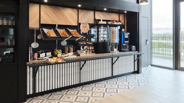 A modern cafe with a counter displaying pastries, a coffee machine, and a refrigerated display. - B&B HOTEL Versailles Rocquencourt (Le Chesnay-Rocquencourt)