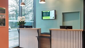 A reception desk with a digital display, a potted plant, and a view of the street outside. - B&B HOTEL Versailles Rocquencourt (Le Chesnay-Rocquencourt)