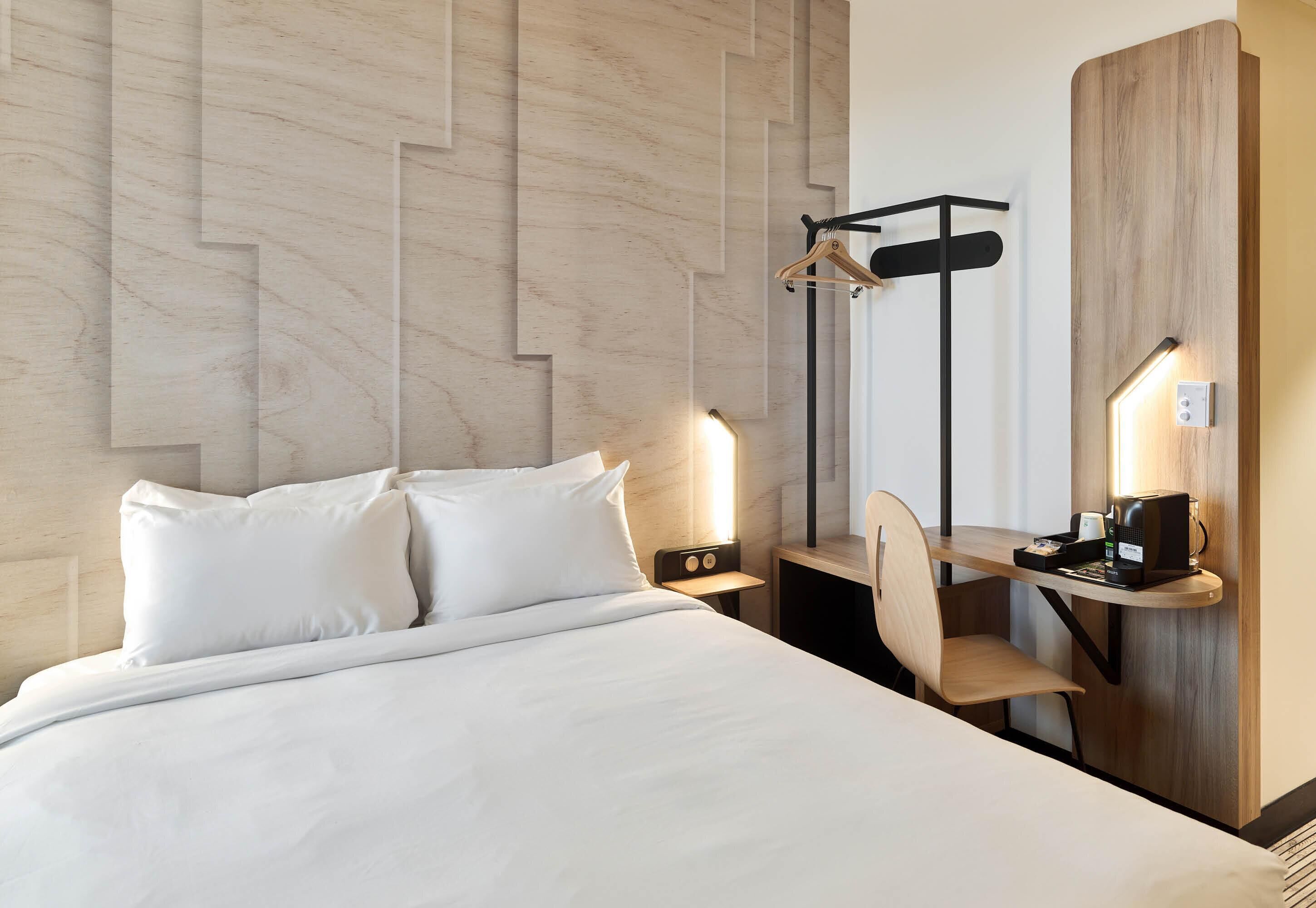 A hotel room with a bed, bedside tables, a desk with a chair, a coat rack, and a wall-mounted light fixture.