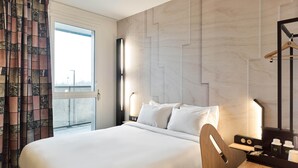A hotel room with a large bed, a desk, a chair, a lamp, and a view of the outside. - B&B HOTEL Versailles Rocquencourt (Le Chesnay-Rocquencourt)