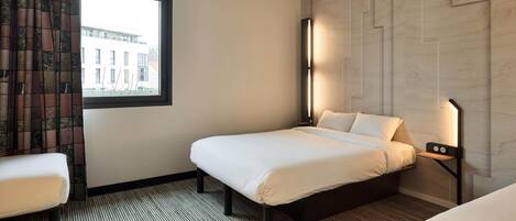 Una habitación de hotel con una cama, una mesita de noche, una ventana con cortinas, una lámpara de pared y vistas a un edificio.