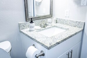 Baño con lavamanos de encimera de granito, espejo, dispensador de jabón, porta cepillos de dientes y porta papel higiénico.