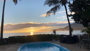 A swimming pool with a view of the sunset over the ocean, surrounded by palm trees and outdoor furniture.