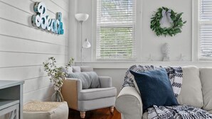 A living room with a light-colored sofa, a small armchair, and a side table with a vase of flowers. There is a wall-mounted sign that reads "Beach" and a wreath on the wall.