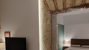 A modern bedroom with a large bed, a television on a wooden stand, and a stone pillar. - Suite in the historic center of Naples (Napoli)