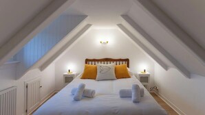 Ein Schlafzimmer mit schrägem Dach, einem Bett mit weißer Bettwäsche und zwei Kissen, einem Nachttisch mit Lampe und einem kleinen Tisch mit einer Tasse.