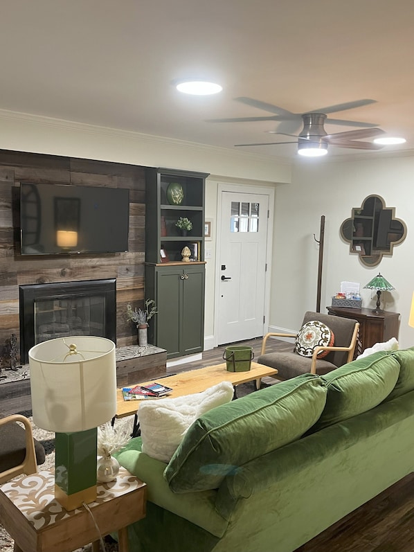 A living room with a green sofa, a coffee table, a TV mounted on the wall, a fireplace, and a ceiling fan. - Family Friendly 3 bedroom close to downtown Raleigh (Raleigh)