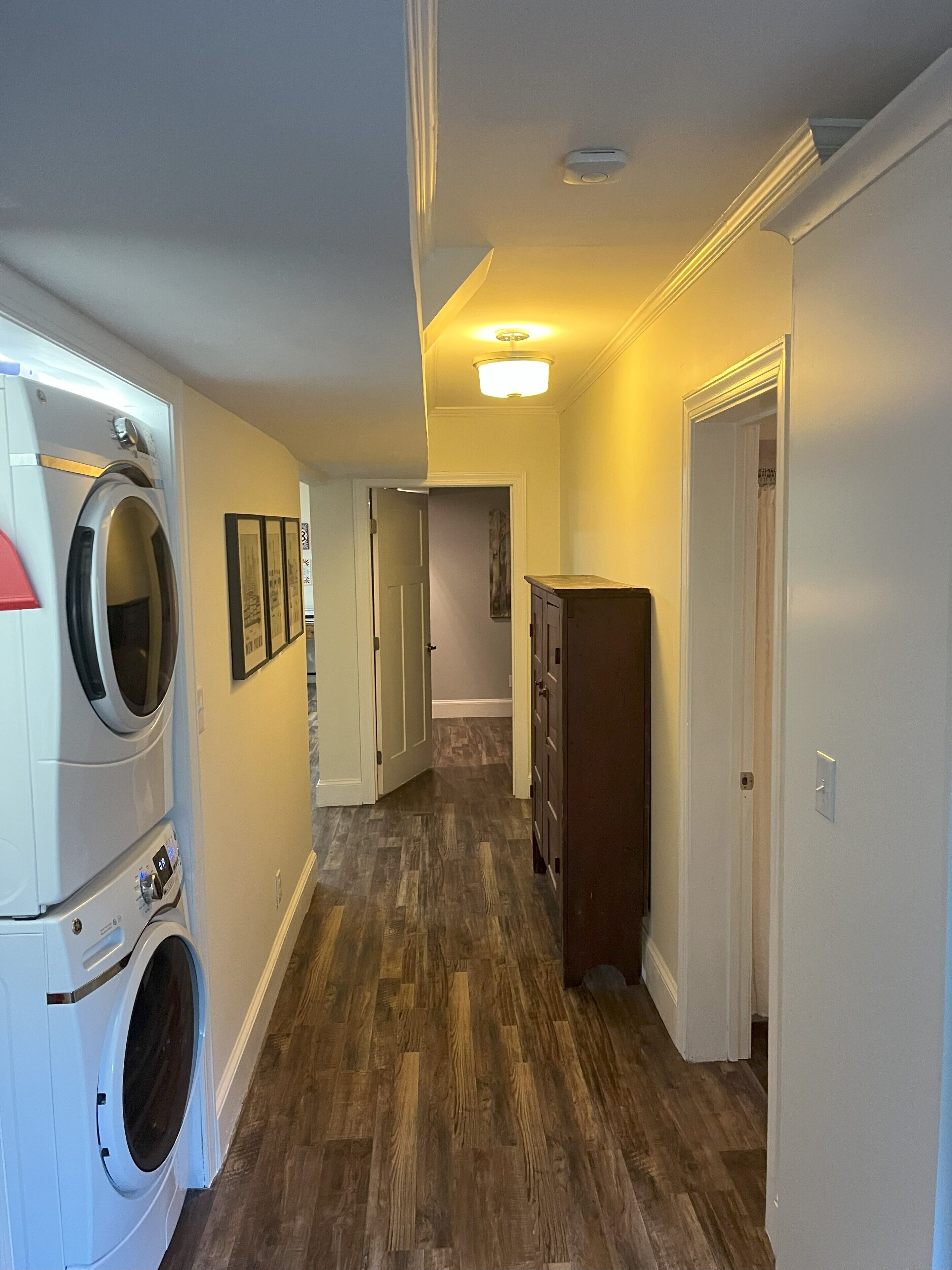 Un cuarto de lavandería con lavadoras y secadoras apiladas, piso de madera y un pasillo que lleva a un dormitorio.