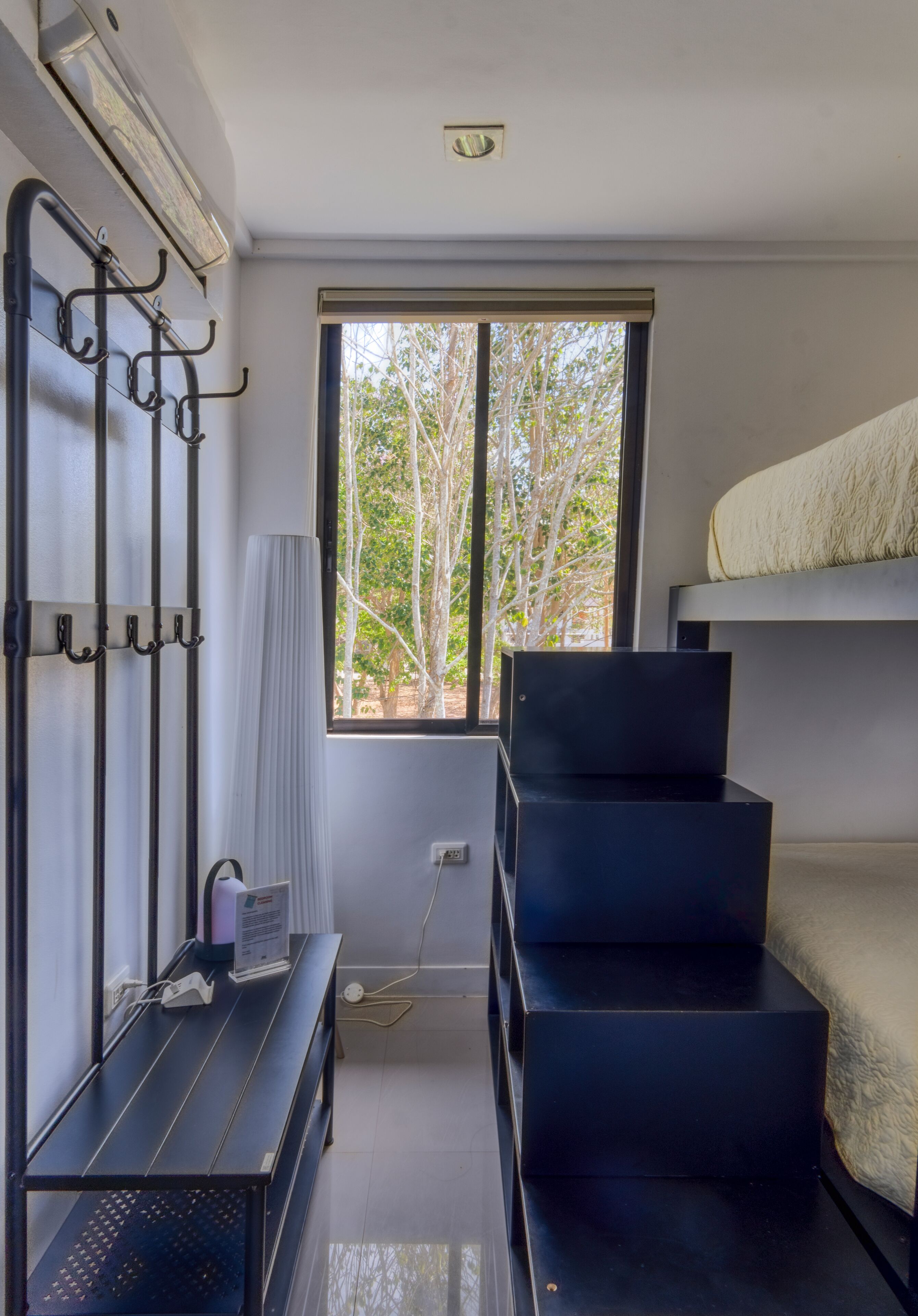 Un dormitorio con literas, un escritorio con computadora, una ventana con vista a árboles y un equipo de aire acondicionado instalado en la pared.