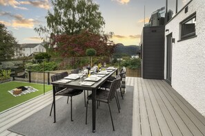 A deck with a dining table set for six, surrounded by chairs, with a view of a house and trees in the background. - NEW - Mayfair House - 5 Star - Lake View Luxury (Bowness-on-Windermere)