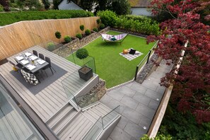A patio with a dining table and chairs, a small grassy area with a hammock, and a wooden deck. - NEW - Mayfair House - 5 Star - Lake View Luxury (Bowness-on-Windermere)