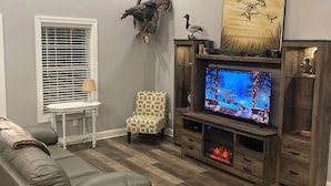 Una sala de estar con un sofá gris, un sillón estampado, un televisor, una chimenea y un ventilador de techo.