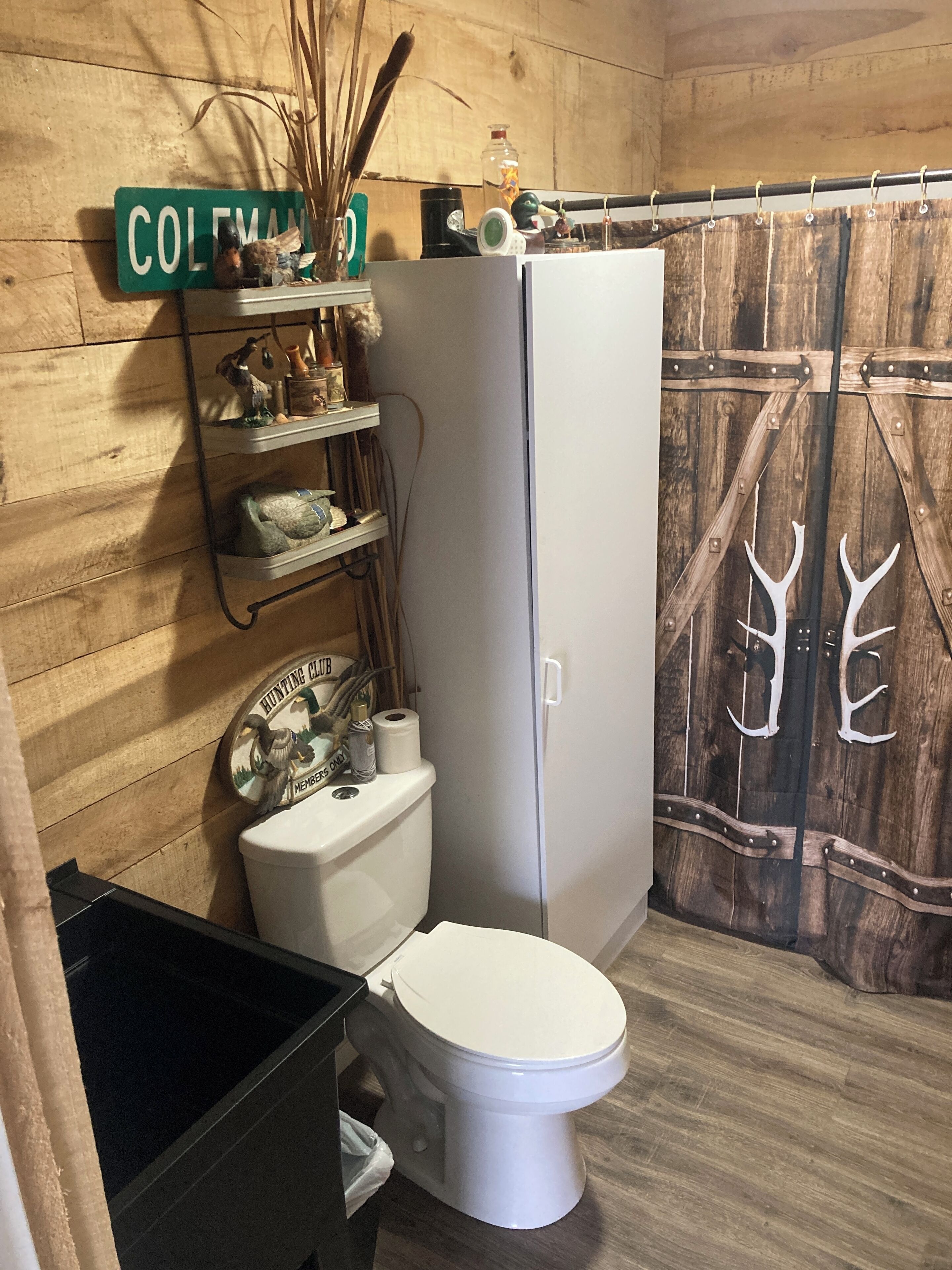 A bathroom with a wooden wall, a toilet, a white cabinet, and a shelf with various items.