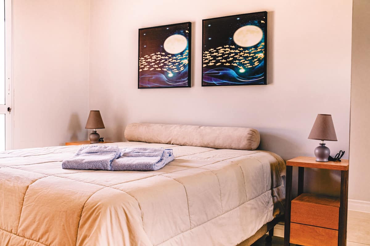 A neatly made bed with a beige duvet, two bedside lamps, and two framed artworks depicting night scenes with moons and stars.