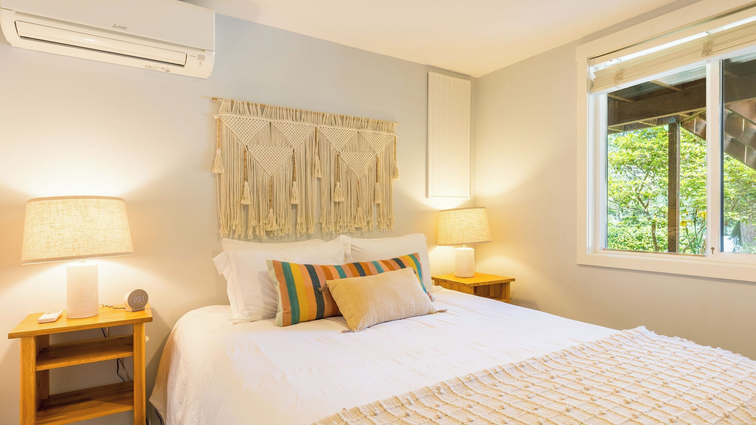 A bedroom with a bed, two bedside tables, a macrame wall hanging, and a window with a view of greenery.