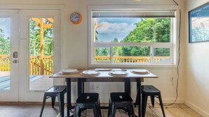 Un comedor con una mesa dispuesta para cuatro personas, una ventana con vistas a árboles y una terraza de madera al exterior.