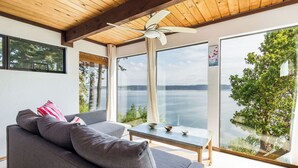 A modern room with a gray sofa, a small table, and a ceiling fan. Large windows offer a view of a lake and trees.