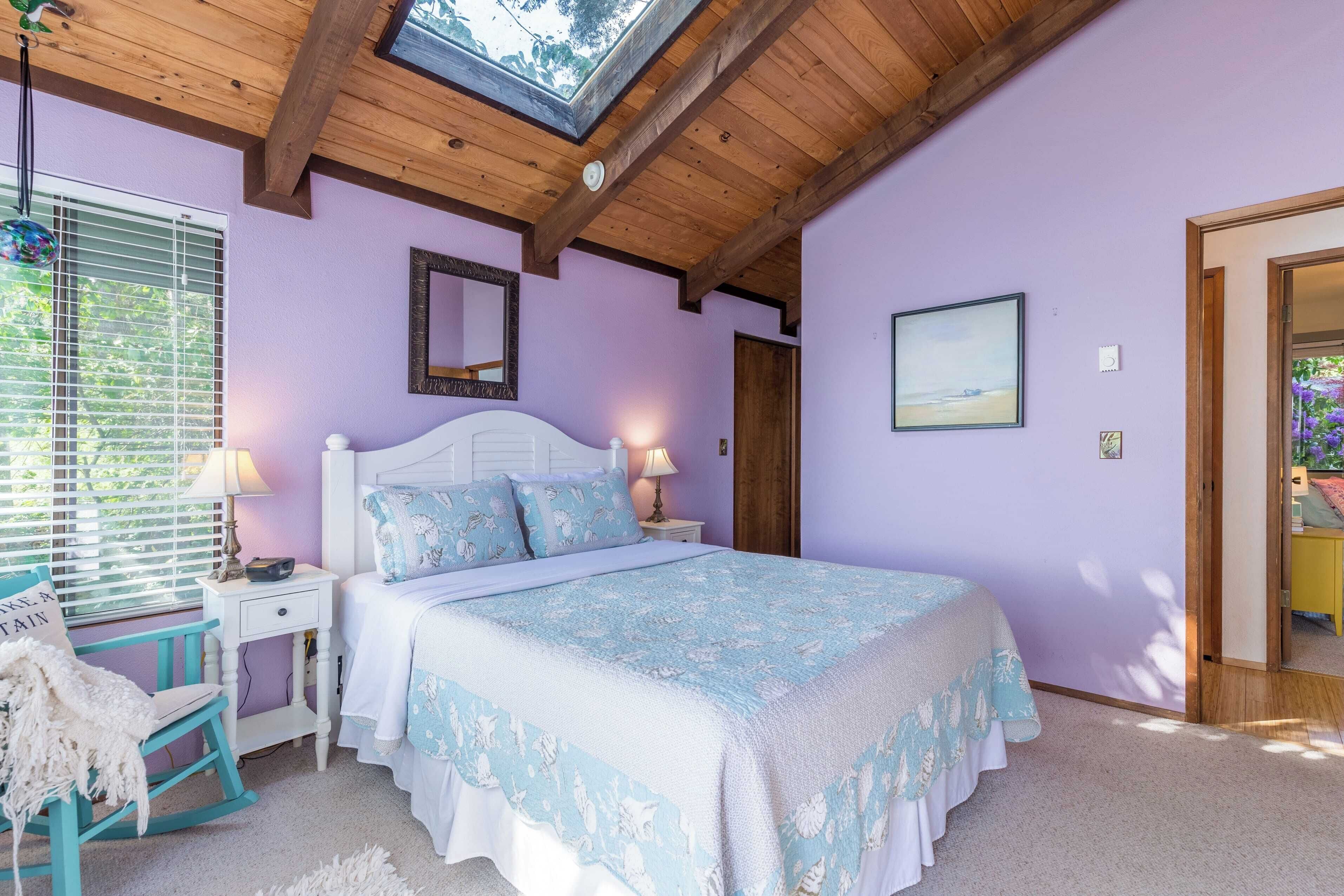 Un dormitorio con techo de madera, una cama con colcha floral, una mesita de noche con lámpara, un sillón mecedor y una ventana con persianas.