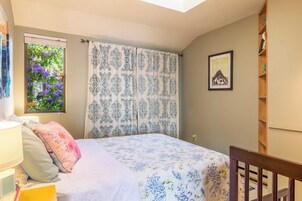 Um quarto com cama, janela com cortinas, mesa de cabeceira, luminária, quadro na parede e uma cadeira de madeira.