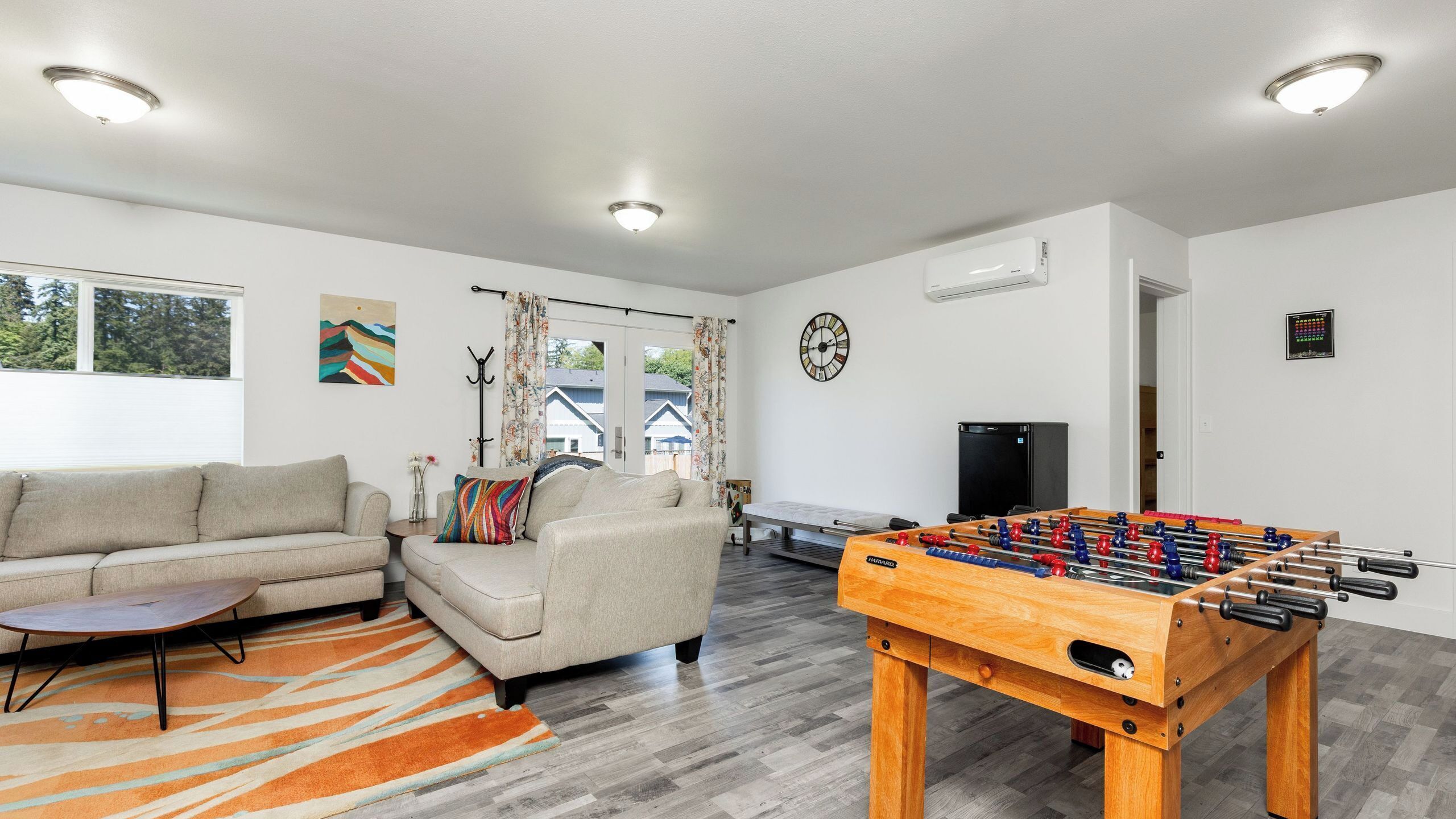 A living room with a sofa, a foosball table, a clock, and a window with a view of houses.