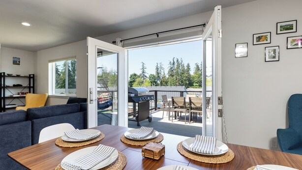 An open sliding door leading to a patio with a grill, a dining table set with plates and napkins, a sofa, and a bookshelf. - Twin Oaks Retreat by Avantstay Mins to Freeland (Freeland)