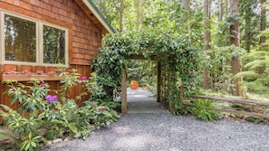 Une cabane en bois, avec un chemin de gravier menant à une arche en bois, elle-même entourée d’une végétation luxuriante.