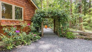 A wooden cabin with a gravel path leading to a wooden archway surrounded by lush greenery. - The Cottage at Herons Roost by Avantstay (Langley)