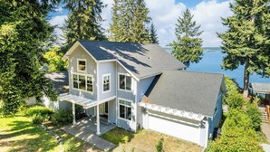 A two-story house with a garage, surrounded by lush greenery and a view of the water. - Panoramic Saratoga Passage Views Elegant Whidbey Home by Avantstay (Langley)