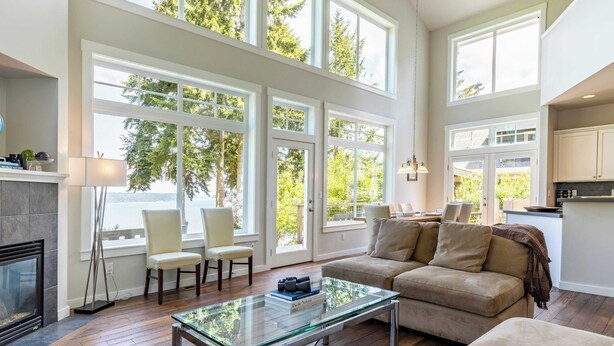 A spacious living room with a glass coffee table, beige sofa, and a fireplace. Large windows provide natural light and views of greenery. - Panoramic Saratoga Passage Views Elegant Whidbey Home by Avantstay (Langley)