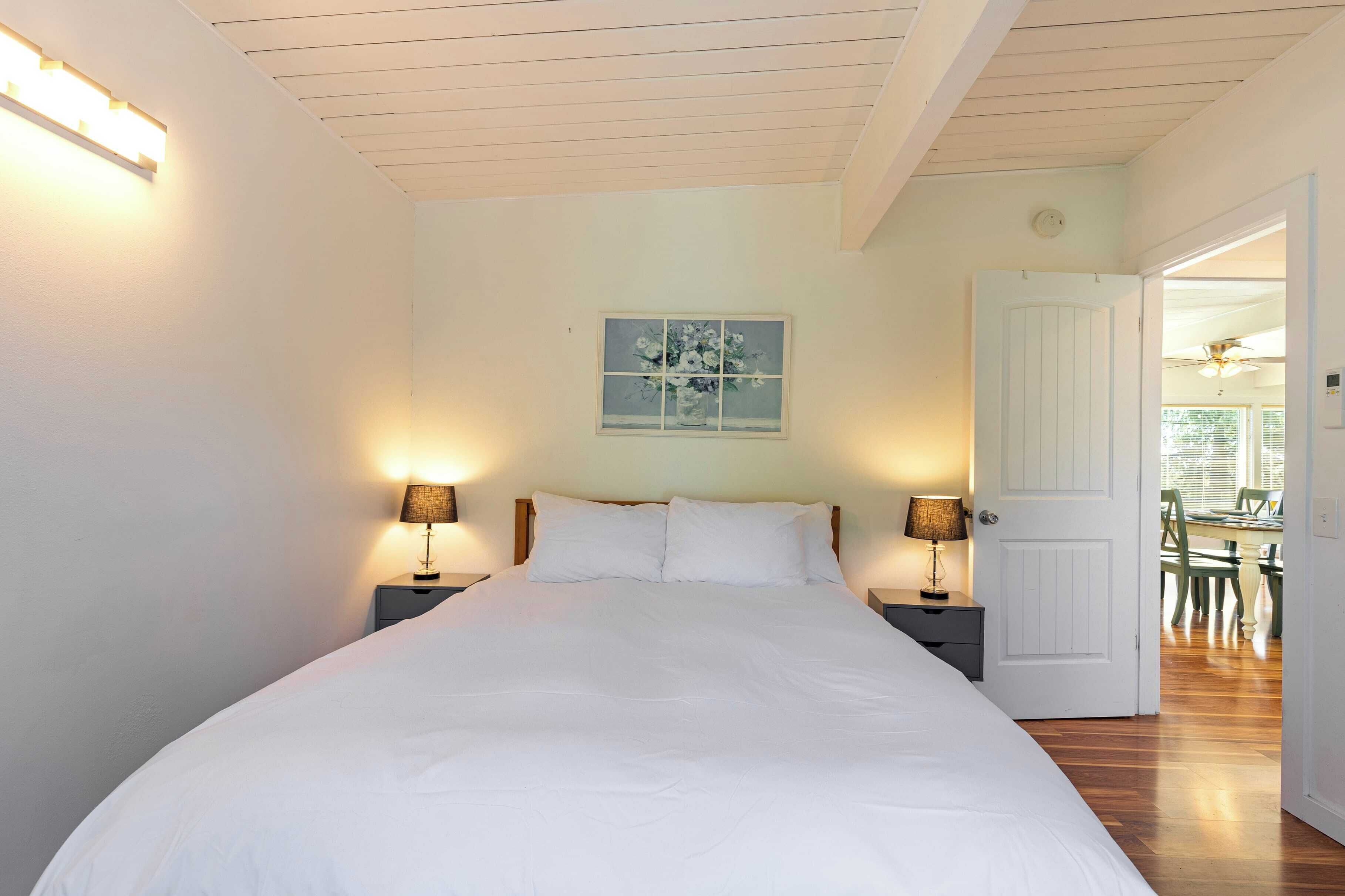A bedroom with a large bed, two bedside tables with lamps, a window with a floral print, and a door leading to another room.