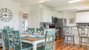 A modern kitchen with white cabinets, a stainless steel refrigerator, and a dining area with teal chairs and a wooden table. - Sunny Side Island Retreat by Avantstay Backyard (Freeland)