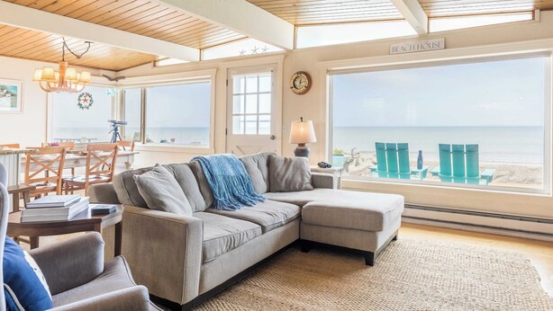 Una sala de estar acogedora con un ventanal grande que da a la playa, techo de madera y un letrero que dice "Beach House".