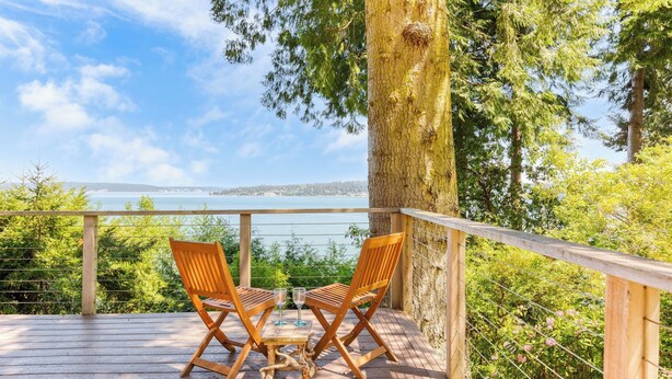 A wooden deck with two wooden chairs and a small table, overlooking a body of water and surrounded by trees. - Serenity Pines by Avantstay Incredible Views (Greenbank)