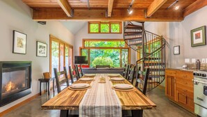 A dining area with a long wooden table set for six, a fireplace, a kitchen with a stove, and a spiral staircase. - Rhythm Waters by Avantstay Garden Paradise, Pond (Langley)