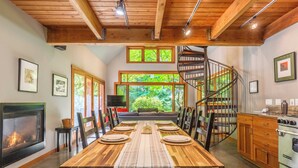 A dining area with a wooden table set for six, a fireplace, and a spiral staircase. - Rhythm Waters by Avantstay Garden Paradise, Pond (Langley)