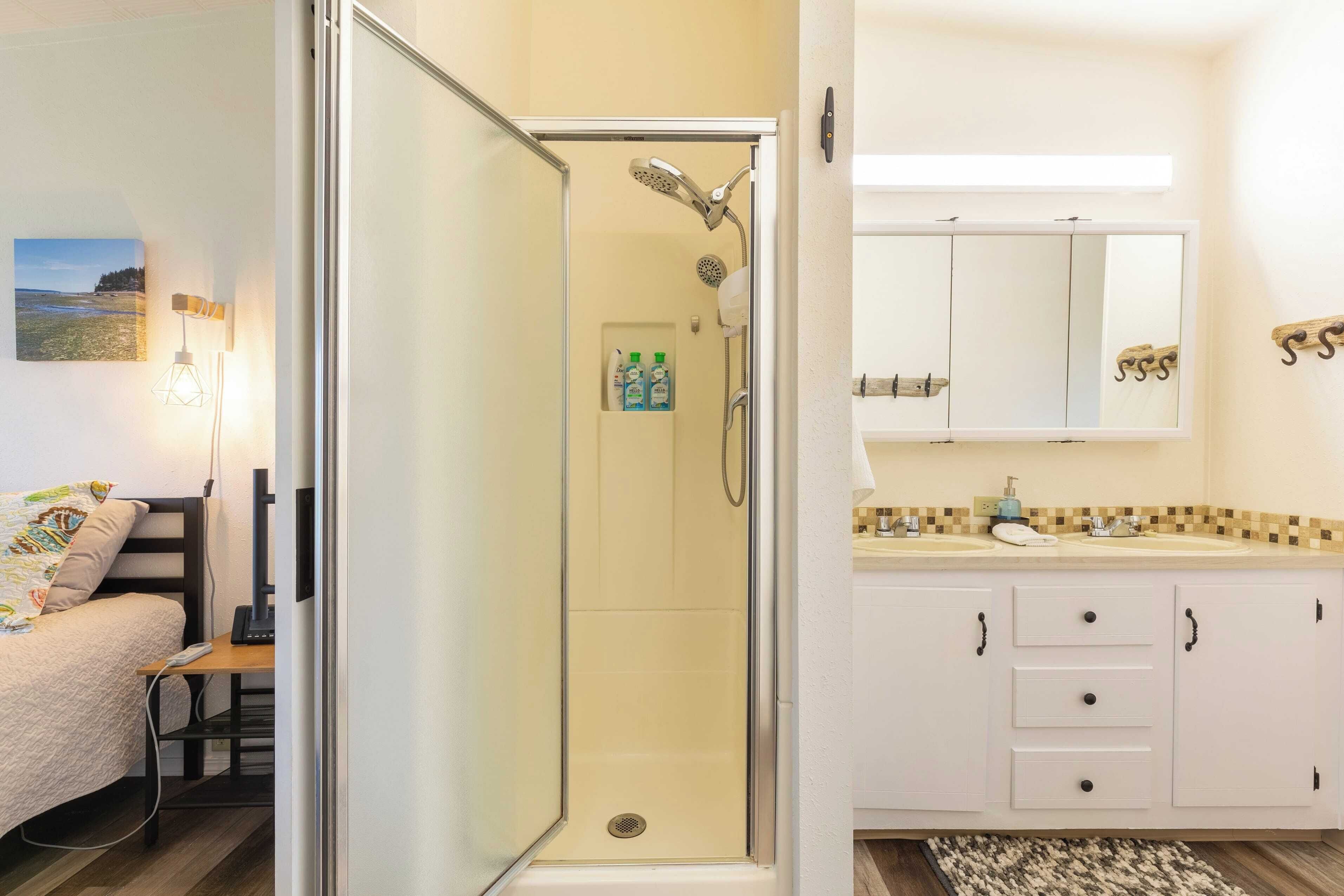 A hotel bathroom with a glass-enclosed shower, a sink with a mirror above it, and a bed with a view of the beach.