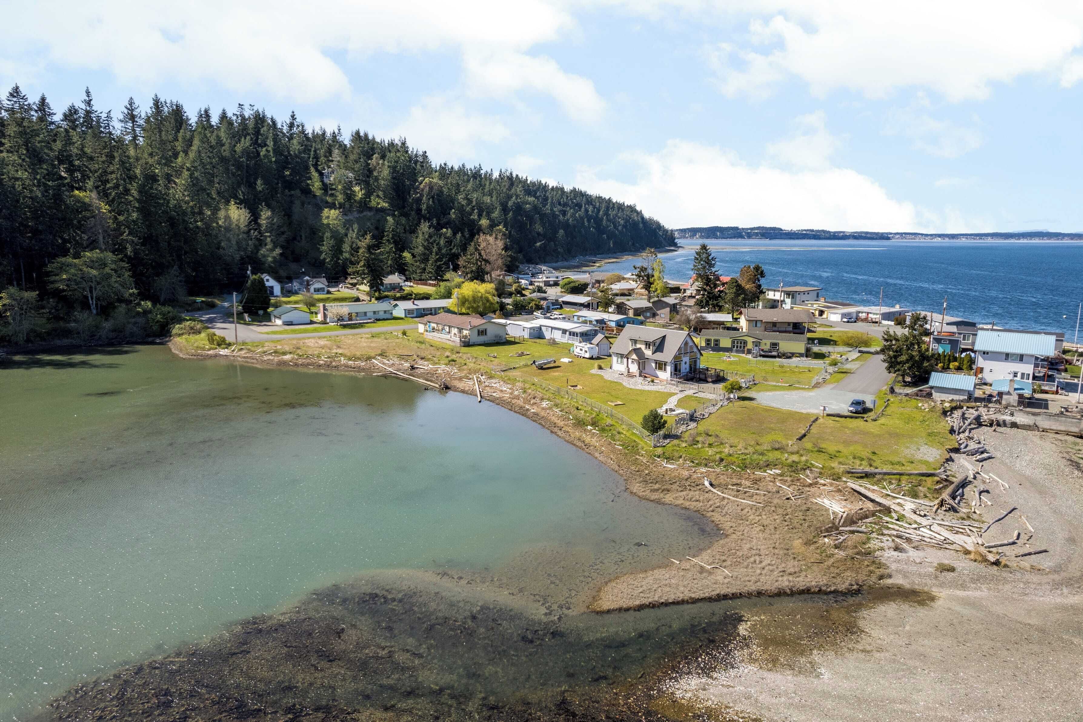 A coastal village with houses along the shoreline, a clear view of the sea, and a forested hill in the background.