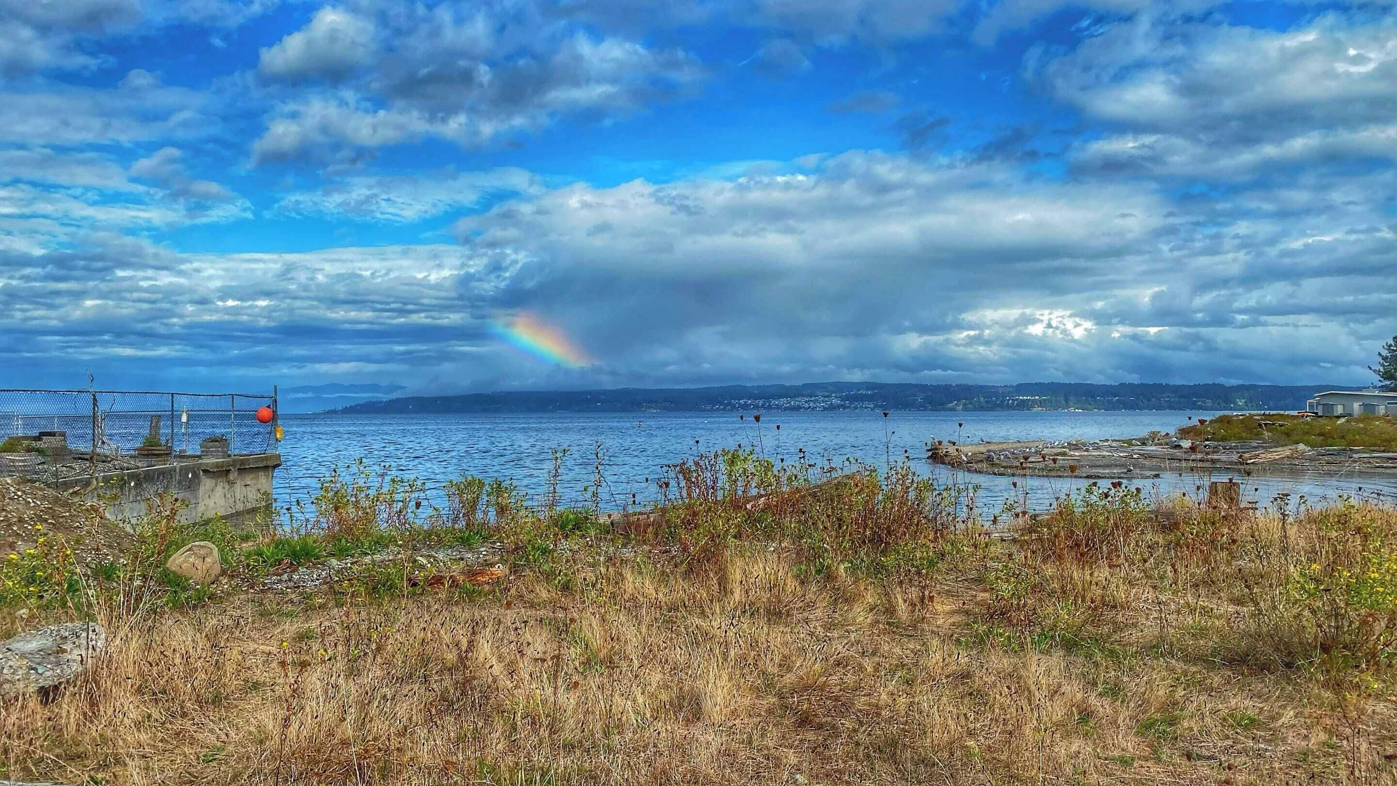 Un paysage côtier avec vue sur la mer, un arc-en-ciel et des collines lointaines.